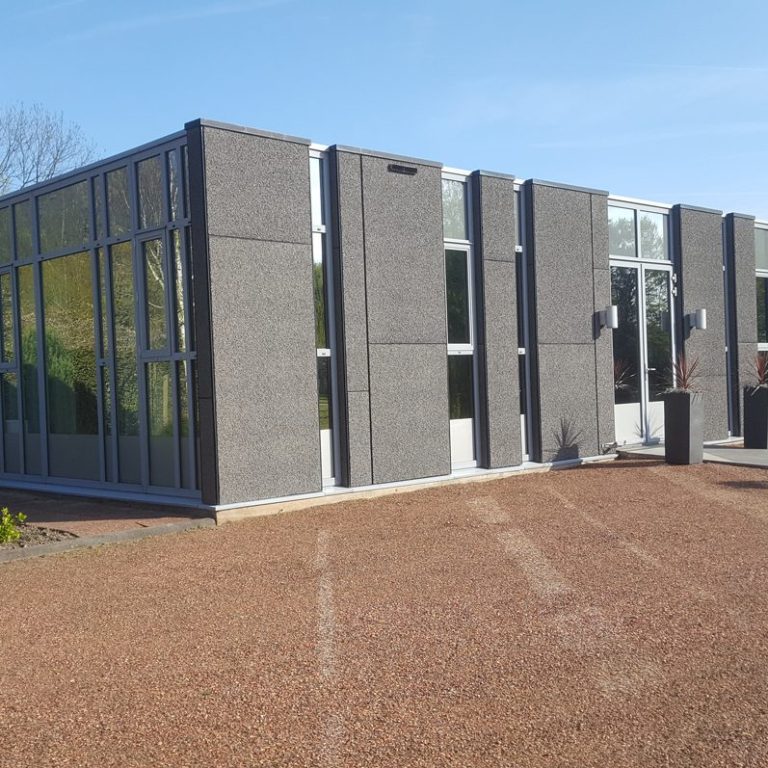 Moderne, vierkante gebouw met grote ramen en grijze gevel, omgeven door gravel.