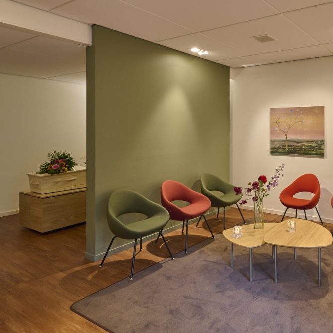 Afscheidsruimte met groene en rode stoelen rond een ronde tafel en een schilderij aan de muur.